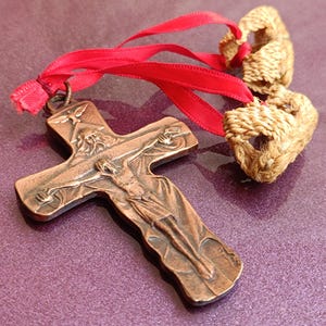 Pode incluir: Uma cruz de cor bronze com uma representação detalhada de Jesus Cristo na cruz, presa a uma fita vermelha e dois ornamentos em forma de coração trançados. A cruz está sobre um fundo roxo.