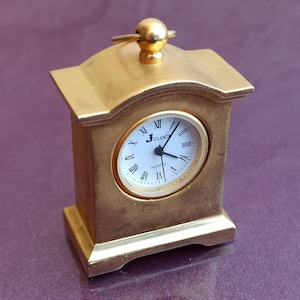 May include: A small, gold-toned, tabletop clock with a white face and black hands. The clock face has Roman numerals and the text "J. CLOCK QUARTZ".