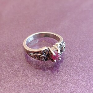 K&ouml;nnte beinhalten: Ein silberner Ring mit einem herzf&ouml;rmigen, tiefroten Edelstein in der Mitte. Der Ring hat dekorative Details am Band und um den Edelstein, vor einem lila Hintergrund.