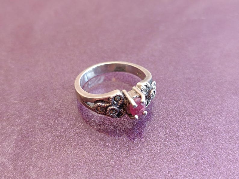 K&ouml;nnte beinhalten: Ein silberner Ring mit einem herzf&ouml;rmigen, tiefroten Edelstein in der Mitte. Der Ring hat dekorative Details am Band und um den Edelstein, vor einem lila Hintergrund.