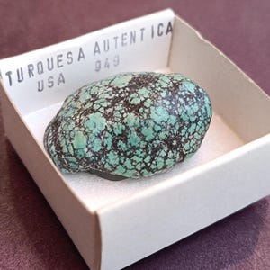 Può includere: Una pietra turchese con un motivo screziato di verde acqua e nero, presentata in una scatola di cartone bianca. La scatola riporta la scritta "TURQUESA AUTENTICA" e "USA". La pietra ha una forma naturale e organica.