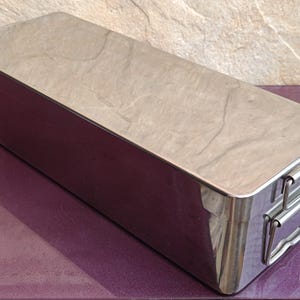 Pode incluir: Uma caixa de armazenamento retangular de aço inoxidável polido com tampa plana e duas alças laterais de metal articuladas. O recipiente apresenta uma superfície metálica limpa e reflexiva com um design seguro e retangular.