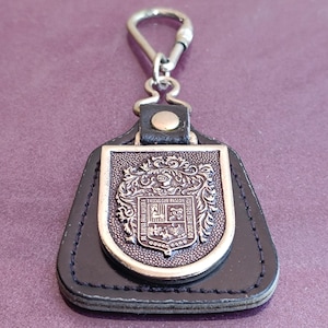 Pode incluir: Um porta-chaves de couro preto com um emblema de metal prateado. O emblema apresenta um design de brasão detalhado com texto e elementos decorativos. O porta-chaves tem um fecho de metal.
