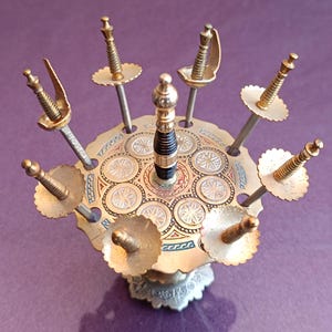 Może przedstawiać: Zestaw siedmiu złotych, ozdobnych uchwytów na wykałaczki w kształcie mieczy, ułożonych w okrągłym stojaku z wzorem w kolorze złotym i białym.