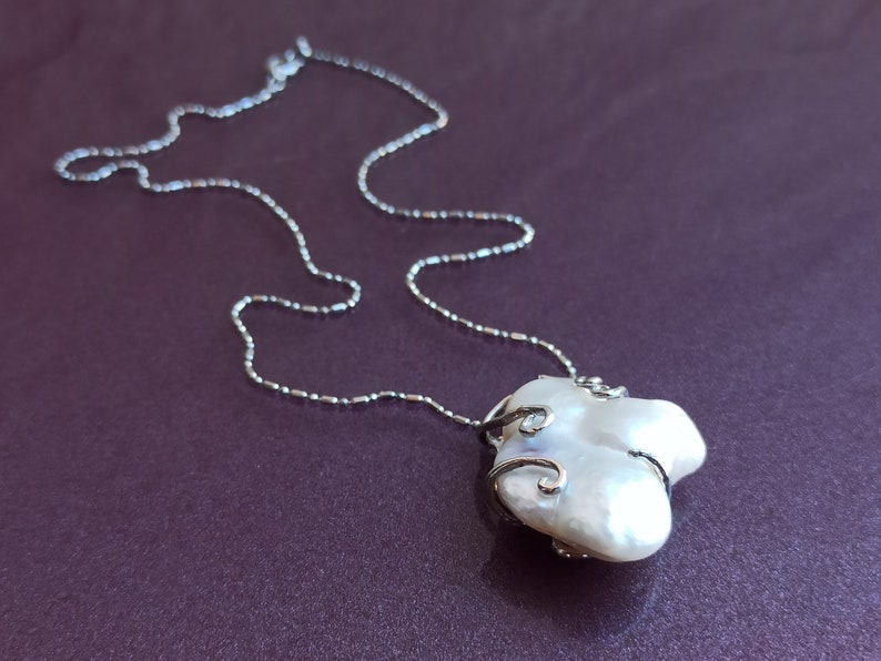May include: A silver chain necklace with a large, white baroque pearl pendant. The pearl is wrapped in silver wire with delicate swirls.