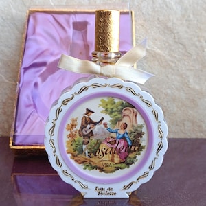 May include: A round glass perfume bottle with a gold cap and a cream ribbon. The front features a decorative scalloped border and a printed scene of a couple in a garden with the text Rosaleda Vida and Eau de Toilette below.