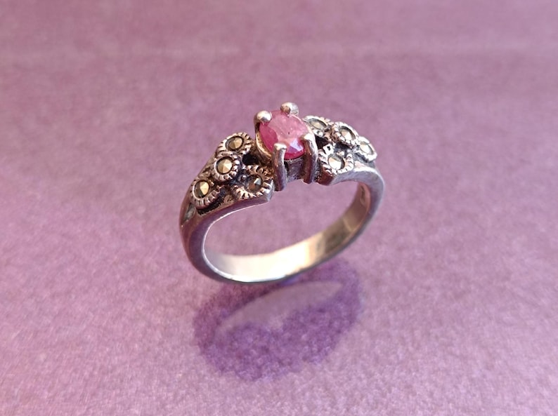 K&ouml;nnte beinhalten: Ein silberner Ring mit einem rosa Edelstein in der Mitte. Der Ring hat ein dekoratives Design mit kleinen, dunklen Akzenten um den Edelstein und am Band. Der Ring ist vor einem lila Hintergrund platziert.
