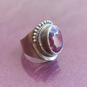 Pode incluir: Um anel de prata com uma grande pedra preciosa rosa de forma oval, facetada, engastada em uma moldura de prata. O anel tem uma banda decorativa com uma borda de contas.
