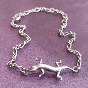 May include: A silver-toned bracelet featuring a lizard charm. The lizard is centered on a chain with a figaro-style link. The bracelet is displayed on a purple surface. The bracelet is a jewelry item.