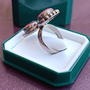 Puede incluir: Un anillo de plata con dos piedras preciosas rosas ovaladas. El anillo est&aacute; engastado en una banda de plata y se muestra en una caja de joyer&iacute;a verde y blanca. Las piedras preciosas son de color rosa vibrante y el anillo tiene un aspecto vintage o antiguo.