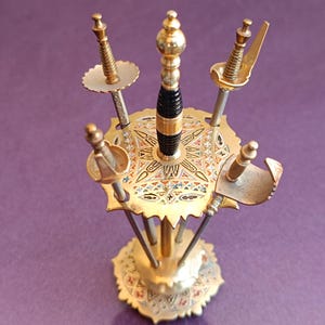 Może przedstawiać: Ozdobny, złoty stojak na miecze z sześcioma miniaturowymi mieczami. Stojak posiada skomplikowane wzory w kolorze czerwonym, niebieskim i białym. Centralny miecz ma czarny uchwyt. Podstawa jest okrągła i również zdobiona.