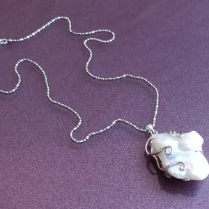 May include: A silver chain necklace with a large, white, irregularly shaped pearl pendant. The pearl is wrapped in silver wire.
