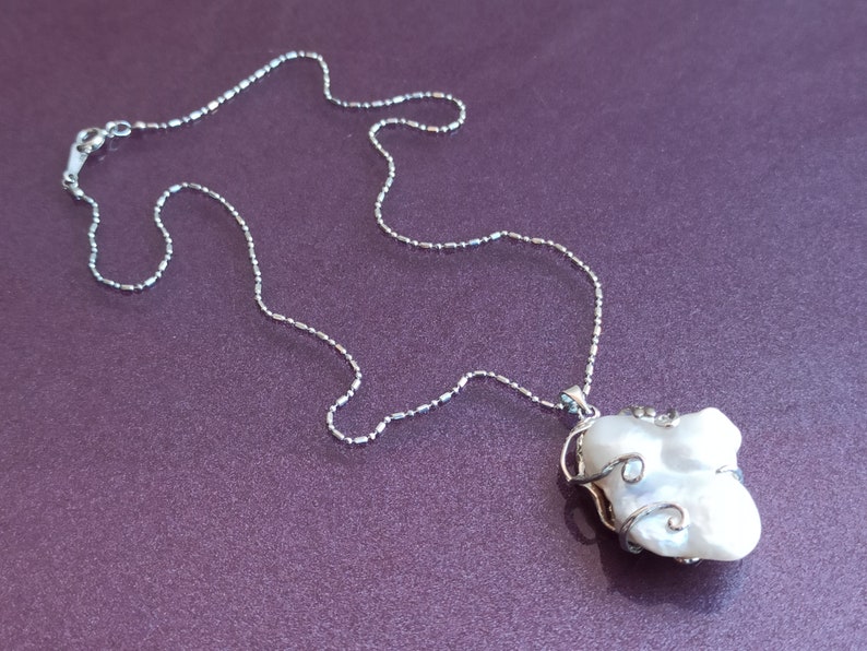 May include: A silver chain necklace with a large, white, irregularly shaped pearl pendant. The pearl is wrapped in silver wire.