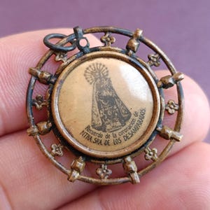 Könnte beinhalten: Ein goldfarbener Metallanhänger mit einem runden Design. Der Anhänger zeigt ein kleines Bild einer Frau in einer Robe mit dem Text "Recuerdo de la coronacion de NTRA. SRA. DE LOS DESAMPARADOS".