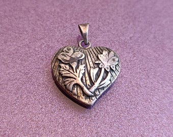 May include: A silver-toned heart-shaped pendant with floral detailing. The pendant features a raised design of a rose and other flowers with leaves. The bail is at the top of the heart. The pendant is on a purple background.