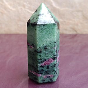 May include: A green and black ruby zoisite crystal obelisk. The polished stone has a hexagonal shape and a pointed top. Pink and red veins run through the green stone. The crystal is approximately 3 inches tall.