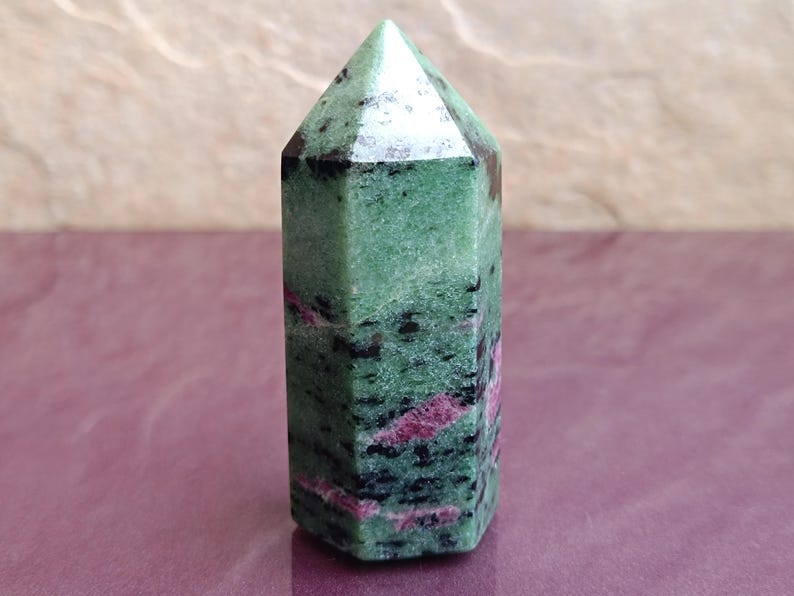May include: A green and black ruby zoisite crystal obelisk. The polished stone has a hexagonal shape and a pointed top. Pink and red veins run through the green stone. The crystal is approximately 3 inches tall.