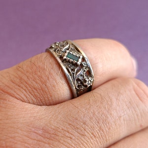 May include: A silver-toned metal ring featuring an intricate, raised floral or vine-like relief pattern across the band. A single, small, rectangular-cut dark green stone is set centrally within the decorative metalwork of the ring band.