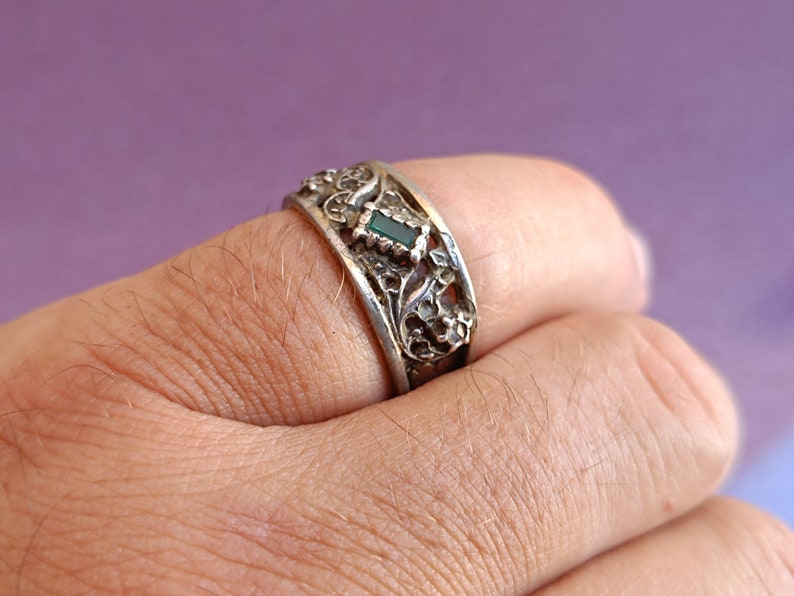 May include: A silver-toned metal ring featuring an intricate, raised floral or vine-like relief pattern across the band. A single, small, rectangular-cut dark green stone is set centrally within the decorative metalwork of the ring band.