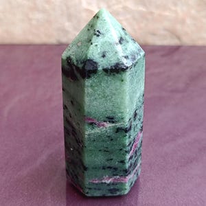 May include: A green and black zoisite crystal point with pink inclusions. The polished gemstone has a hexagonal shape and a pointed top. The crystal is displayed on a purple surface.