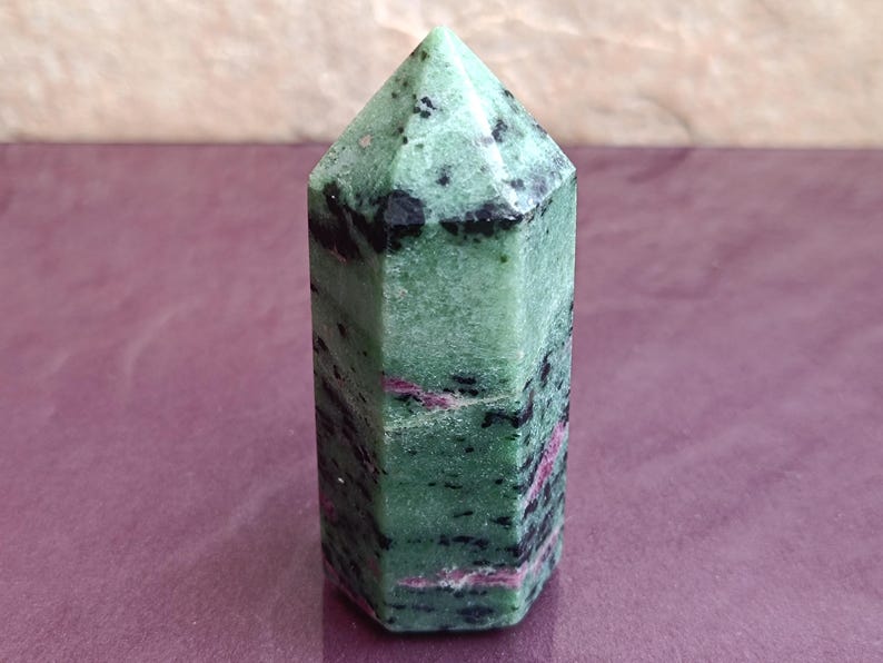 May include: A green and black zoisite crystal point with pink inclusions. The polished gemstone has a hexagonal shape and a pointed top. The crystal is displayed on a purple surface.
