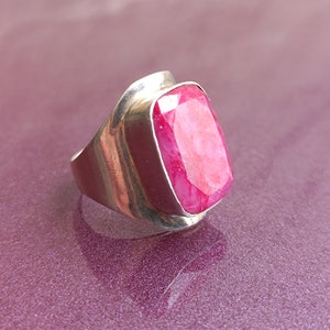 May include: A silver ring with a large, rectangular, faceted ruby gemstone. The ruby is a vibrant pink-red colour and is set in a smooth, polished silver band. The ring has a modern, bold design.