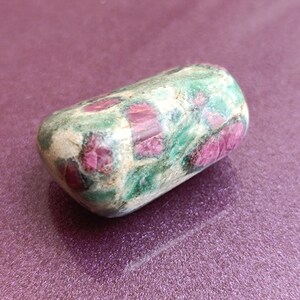 May include: A rounded, tumbled gemstone. The stone has a light green base with red and pink inclusions. The surface is smooth and polished. The gemstone is likely a ruby. The stone's shape is approximately 5 cm long.