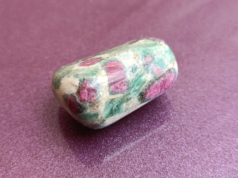 May include: A rounded, tumbled gemstone. The stone has a light green base with red and pink inclusions. The surface is smooth and polished. The gemstone is likely a ruby. The stone's shape is approximately 5 cm long.
