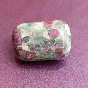 May include: A polished, rectangular gemstone with a mottled appearance. The stone features a base of green and tan hues, speckled with vibrant red ruby inclusions. The gemstone is set against a purple background.
