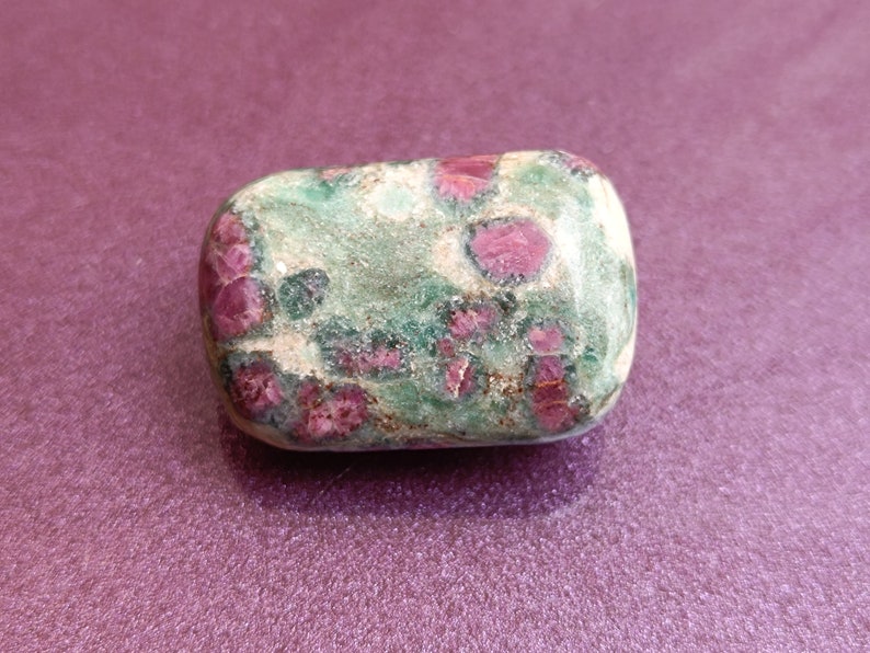 May include: A polished, rectangular gemstone with a mottled appearance. The stone features a base of green and tan hues, speckled with vibrant red ruby inclusions. The gemstone is set against a purple background.