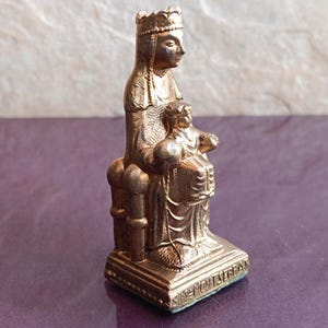 Puede incluir: Estatua dorada de una figura sentada sosteniendo otra más pequeña. La estatua, con corona, se apoya en una base rectangular con texto. Probablemente un artefacto religioso o histórico.