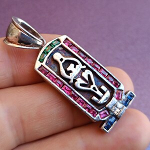 May include: Silver pendant with a detailed design of an Egyptian symbol surrounded by pink, red, and green gemstones. The pendant has a loop at the top for attaching to a chain.