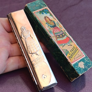 May include: A silver-toned metal harmonica featuring an embossed figure and the text O FADO PORTUGUEZ, accompanied by a rectangular green cardboard box with a colourful illustration of a person in traditional attire and the text O FADO PORTUGUEZ.