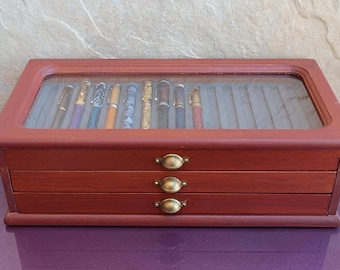 May include: A wooden box with a glass top for storing pens. The box has three drawers and brass handles. The box is a dark reddish brown color.