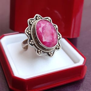 Op de afbeelding: Een zilveren ring met een grote, ovale robijn edelsteen. De robijn is felroze en gezet in een decoratieve bezel met een kralenrand. De ring wordt gepresenteerd in een rode sieradendoos.