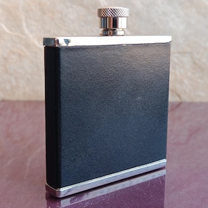 May include: A rectangular, black flask with a textured surface and a silver trim. The flask has a silver cap with a ridged design. The product is designed to hold liquids.