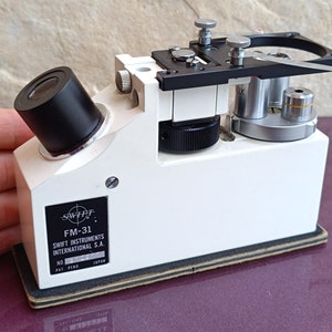 May include: White microscope with a black eyepiece and a black knob. The microscope has a label that reads "SWIFT FM-31 SWIFT INSTRUMENTS INTERNATIONAL S.A. NO. PAT. PEND. JAPAN".