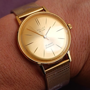 Peut inclure: Une montre Poljot de Luxe dorée avec un bracelet en maille. Le cadran de la montre est doré avec des aiguilles et des marqueurs noirs. Le texte "23 jewels shockproof" est visible. La montre est sur un poignet.