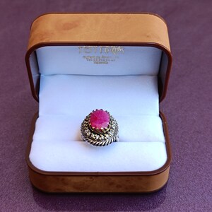 Op de afbeelding: Een ring met een grote ovale roze edelsteen, omringd door kleinere witte edelstenen. De ring ligt in een bruine fluwelen ringdoos.