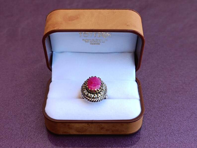 Op de afbeelding: Een ring met een grote ovale roze edelsteen, omringd door kleinere witte edelstenen. De ring ligt in een bruine fluwelen ringdoos.