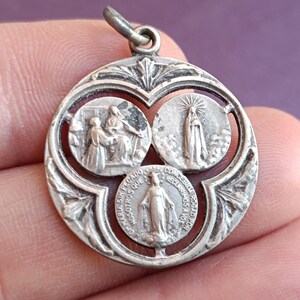 May include: A silver-toned religious pendant with three circular images. The top two depict figures, and the bottom one shows a standing figure with text around the edge. The pendant has a decorative openwork design.