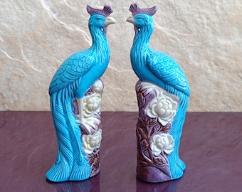 Könnte beinhalten: Zwei blaue Keramikpfauen mit weißen und violetten Akzenten stehen auf einem Sockel mit weißen Blumen. Die Pfauen haben detaillierte Federn und sind ein traditionelles chinesisches Design.