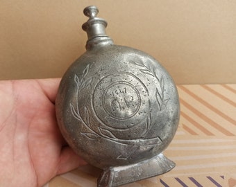 Könnte beinhalten: Eine silberne Metallflasche mit einem verzierten Design, das einen Kranz und die Inschrift "1817" zeigt.