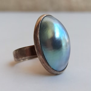 May include: A silver ring with a large, oval-shaped, blue-green pearl.