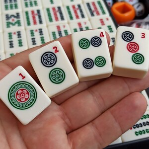 Może przedstawiać: Cztery białe kostki mahjong z czarnymi i zielonymi symbolami i czerwonymi kropkami. Kostki są ponumerowane od 1 do 4.