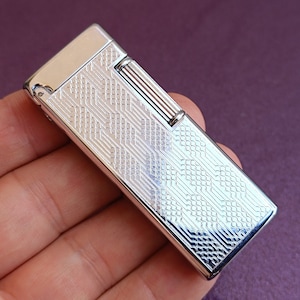 Op de afbeelding: Een zilveren aansteker met een geometrisch patroon aan de voorkant. De aansteker bevindt zich in een hand.