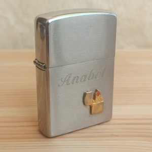 K&ouml;nnte beinhalten: Silberner Feuerzeug mit dem Namen Anabel, der auf der Vorderseite eingraviert ist. Das Feuerzeug hat ein kleines goldenes Emblem mit einem Flammenmotiv.