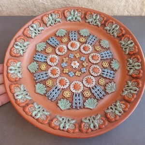 May include: A terracotta plate with a decorative design of blue, green, and yellow flowers and swirls. The plate has a raised rim and a smooth surface.