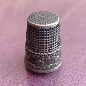 Op de afbeelding: Een zilveren vingerhoed met een decoratief patroon van kronen en bladeren. De vingerhoed heeft een getextureerd oppervlak en een gladde, afgeronde bovenkant.