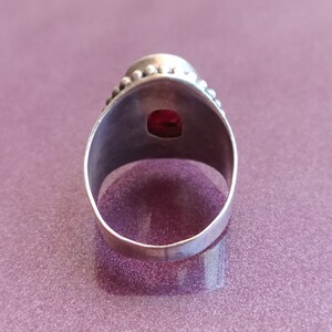 Pode incluir: Um anel de prata com uma pedra preciosa vermelha engastada no centro. O anel tem uma banda com contas.